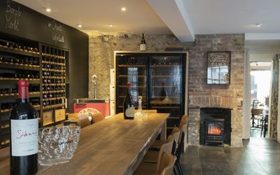 A long wooden banquet table is in a stone room with wine racks and fridges along one wall.