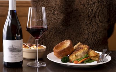 A dark wood table has on it a plate of Sunday lunch, a bottle and glass of red wine and a side of cauliflower cheese