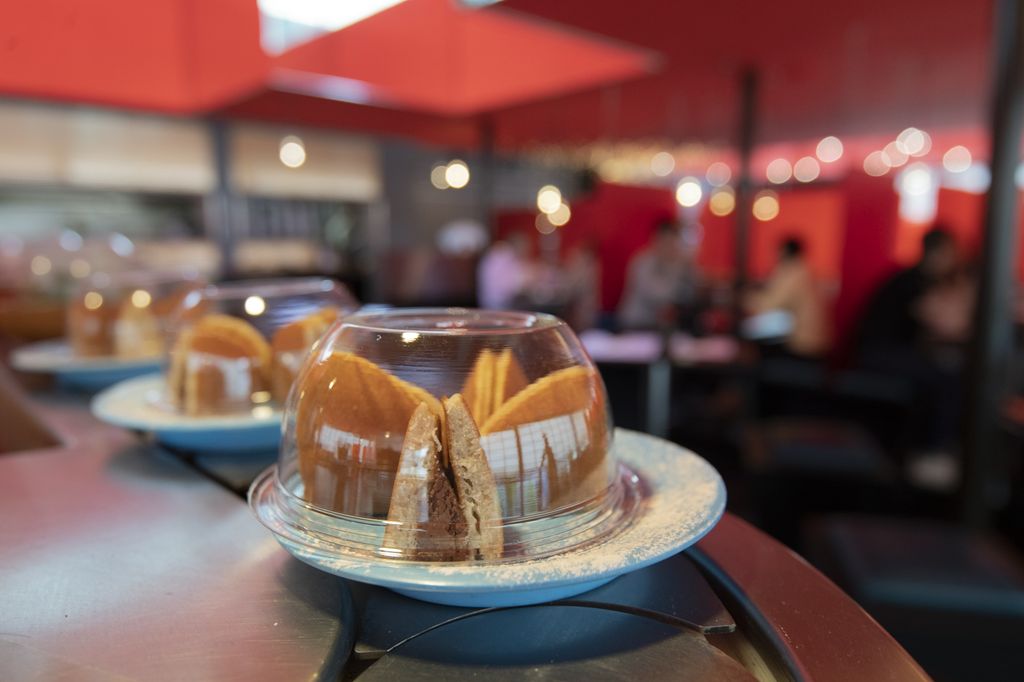 Sushi on a conveyor belt under a plastic dome, a red restaurant in the background.