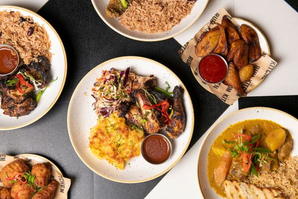 table laid out with caribbean dishes including plantain with red sauce, chicken wings, mashed potatos, jerk chicken and rice