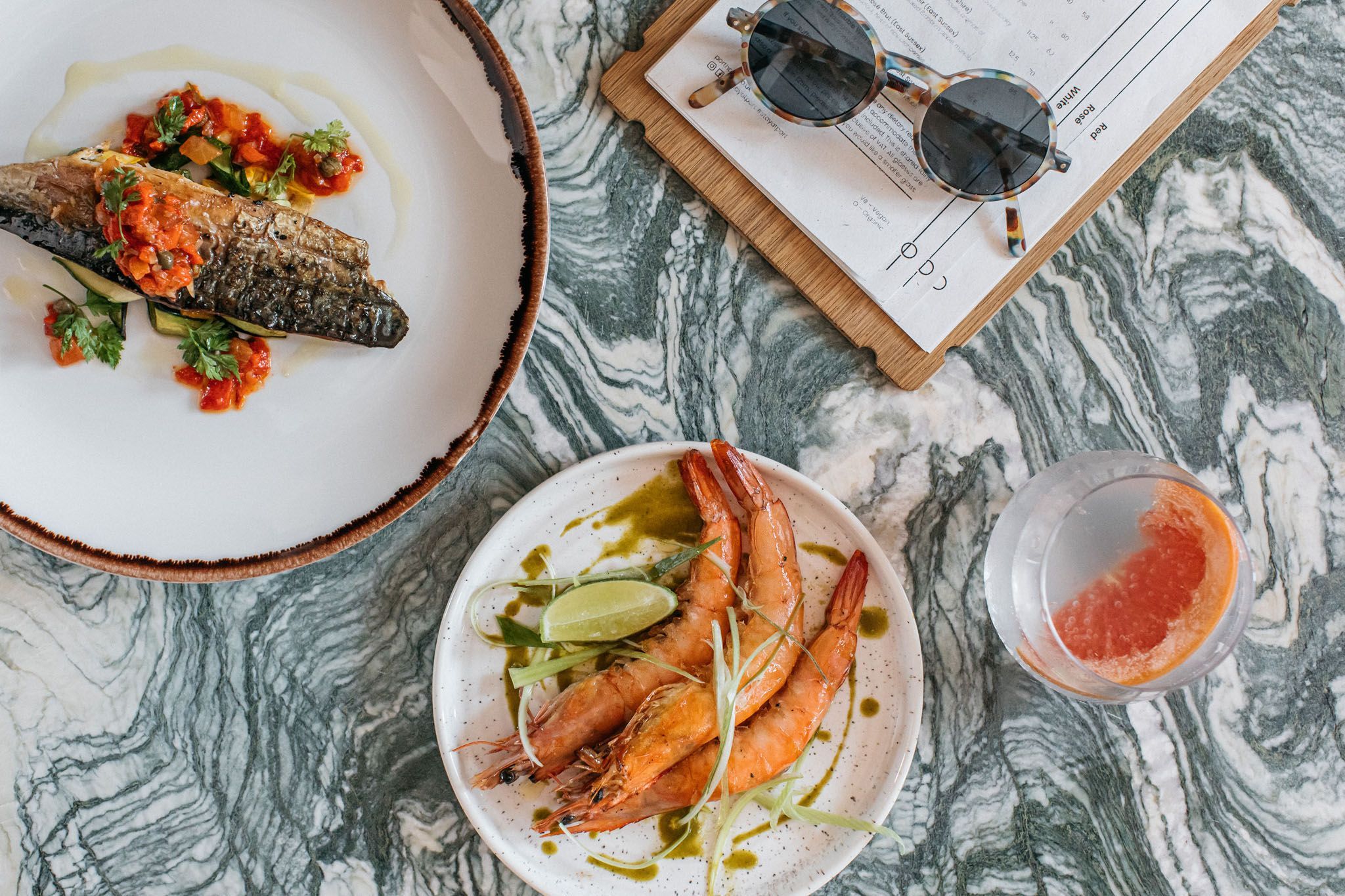 overhead shot of table laid out with small seafood plates including shrimps, glass of wine, menu book, sunglasses