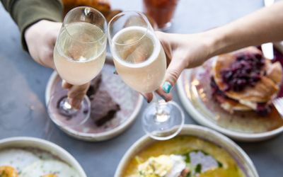 two glasses of prosecco being cheersed over plates of brunch including pancake and eggs
