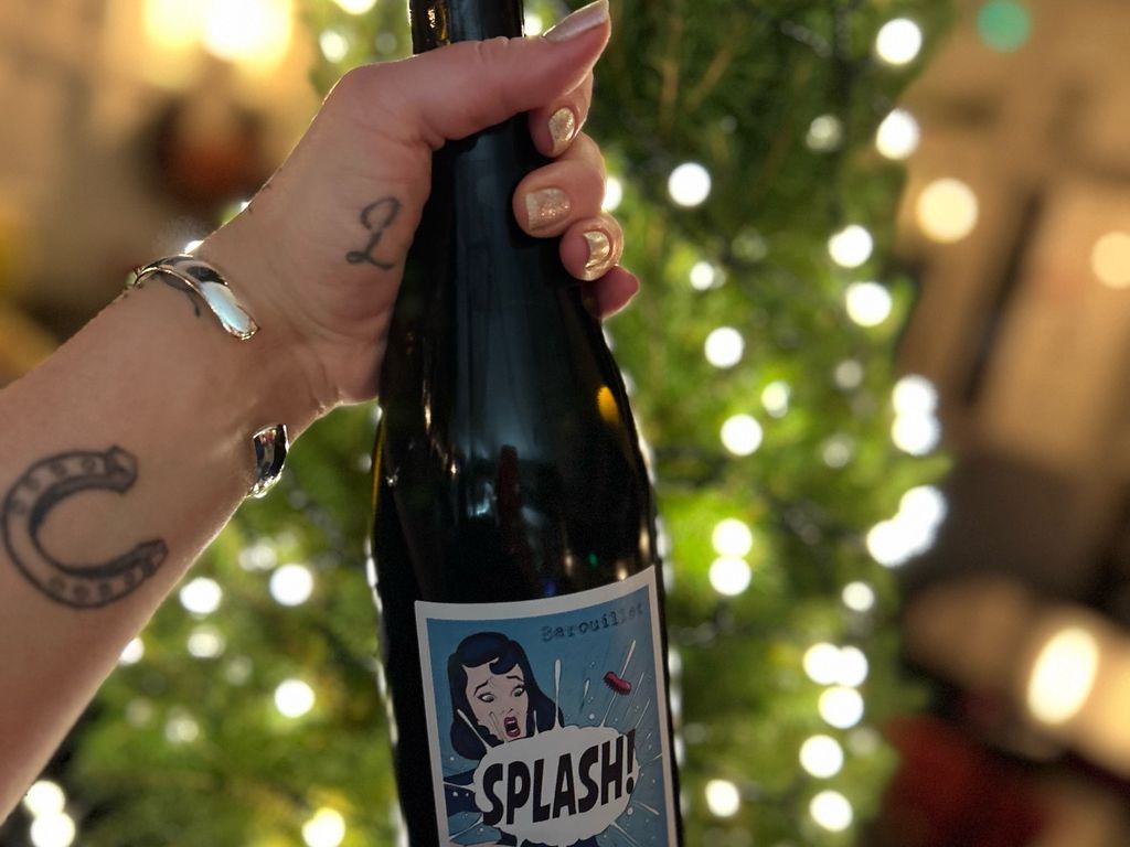 A hand holding up the neck of a wine bottle in front of a Christmas tree, the label reads "Splash!"
