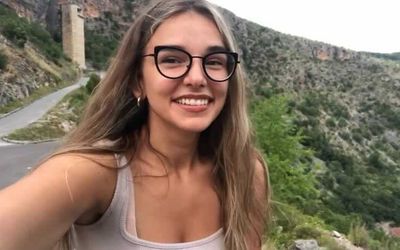 Image of a woman in her 20s, photographed with karst hills in the background. She wears glasses and has long brown hair. She is smiling.