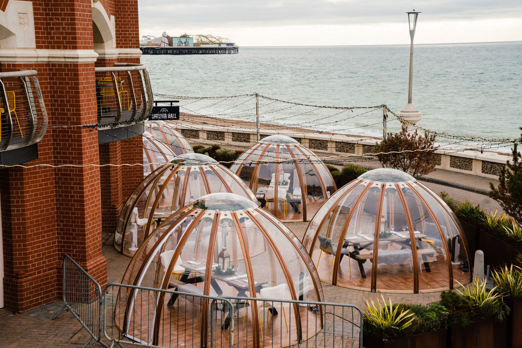 Brighton seafront restaurants. A picture of the Brighton igloos next to Shelter Hall and the Brighton Pier in the background. The igloos are clear in appearance with what seems like wooden struts. They are next to a red brick wall. It is an overcast day.