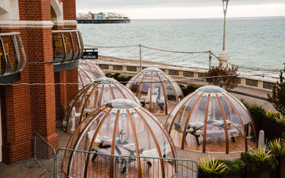 Brighton seafront restaurants. A picture of the Brighton igloos next to Shelter Hall and the Brighton Pier in the background. The igloos are clear in appearance with what seems like wooden struts. They are next to a red brick wall. It is an overcast day.