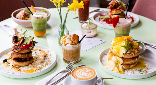 side shot of the light green table laid out with four brunch dishes including frumpets, eggs on the toast, veggie bowl, coffes and smoothes
