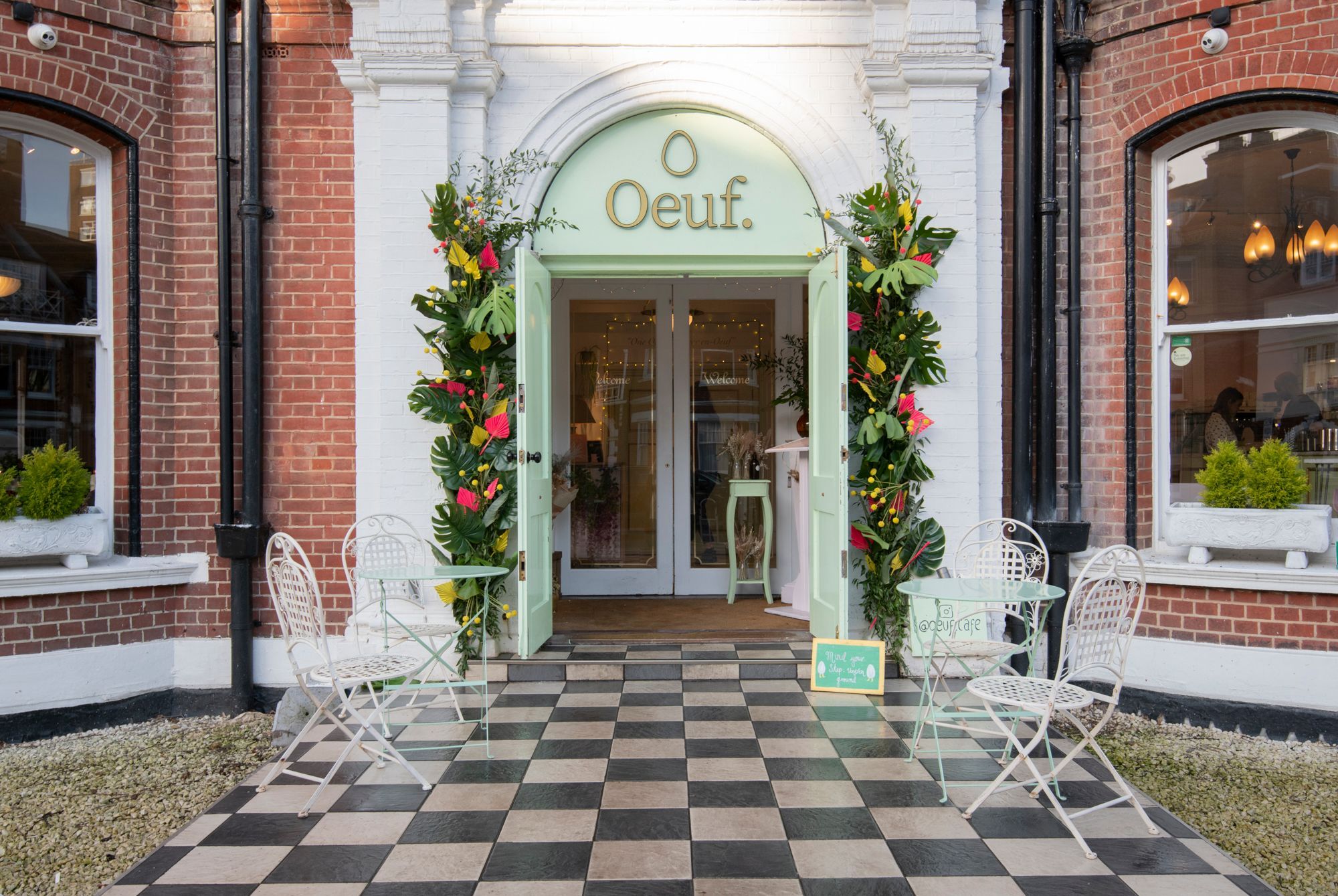 The entrance to Oeuf Cafe, Hove, a black and white tiled walkway with white metal tables and chairs flanking it. The entrance is an egg shell green door with archway and the name Oeuf in gold and a floral arch. White pillars on either side. Flanking that is a red brick facade and bay large bay windows