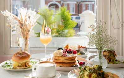 A table by a window laid with brunch, including coffee mimosa, pancakes and burgers. All items available for Brighton takeaway or home delivery.