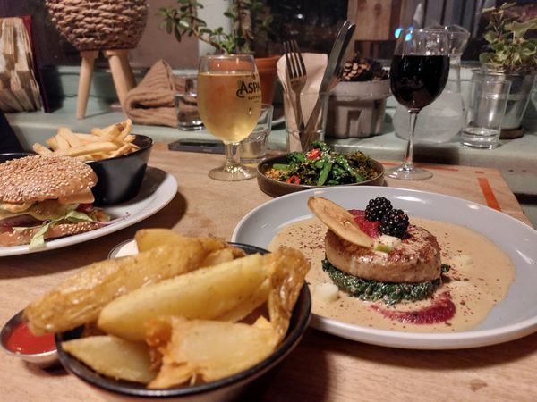 A wooden table with food on it, including potato wedges, a celeriac dish, a burger a pint of cider and a glass of red wine