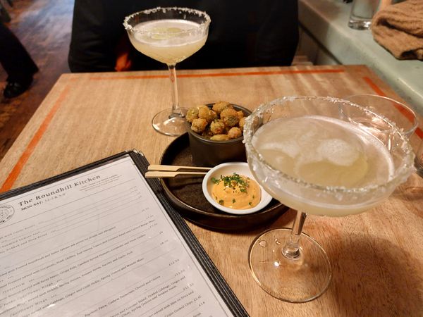 margarita cocktails and panko breaded olives on a wooden table with a food menu