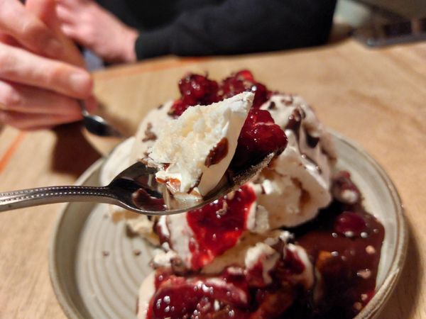 Close up of a spoon with pavlova and berry compote on it and the rest of the pavlova is in the background on a wooden table