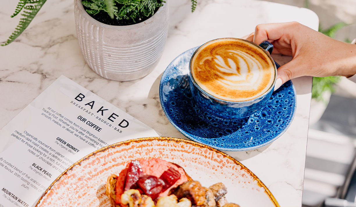 hand holding cup of coffee, there is Baked menu and pancake on the table as well