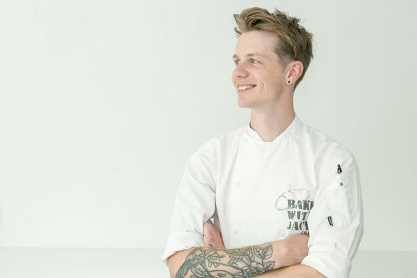 A young man with blond spiked and messy hair is smiling and looking right, he's waring chef whites and has tattoos over his right arm