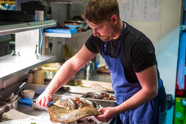 A chef in a black t-shirt and blue apron is in a kitchen preparing a fish dish