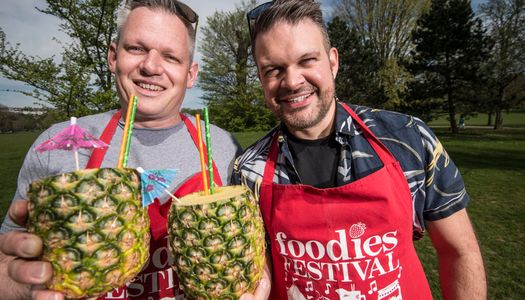 Two men (Duncan Ray and Kenny Tutt) stand side by side in a park holding pineapples with straws in, they're both wearing red Foodies Festival aprons and have sunglasses on their heads