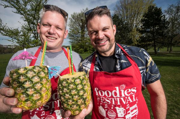 Two men (Duncan Ray and Kenny Tutt) stand side by side in a park holding pineapples with straws in, they're both wearing red Foodies Festival aprons and have sunglasses on their heads