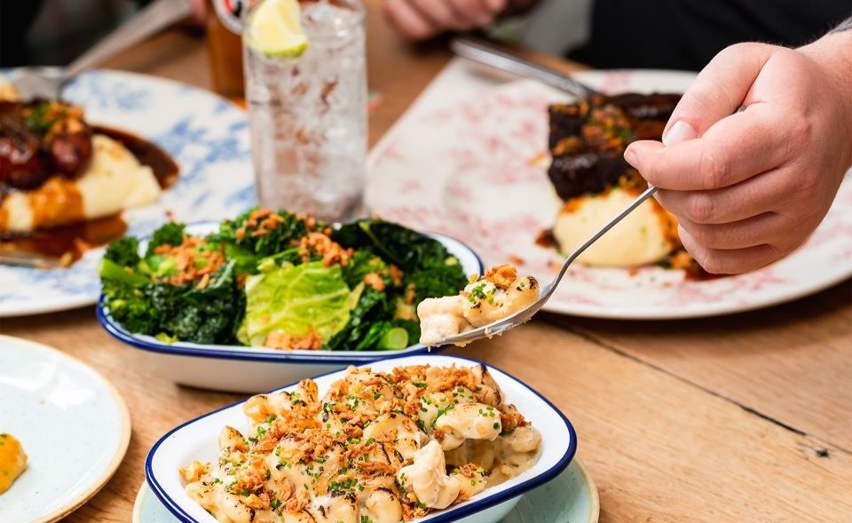side shot of person taking some food from bowl. there are mash potatoes with sausage on the table as well as bowl of broccoli