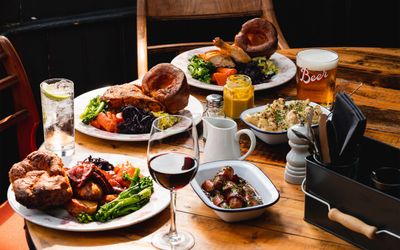 side shot of the table with the three delicious plates of Sunday roast. There are some spices, broccoli and bread rolls on the table as well as glass of beer and glass of red wine