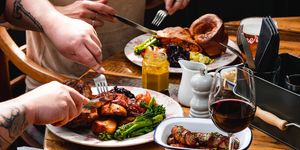 two people eating sunday roast on the brown table, there is gravy in cup, glass of wine, yellowish sauce and some spices on the table too