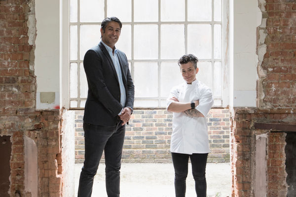 A man in a dark blazer and blue shirt stands next to a female chef in chef whites with short dark curly hair. They are in a building that is light with white morat and exposed brinks that is in the process of renovation. A large window is behind them filling the room with light.