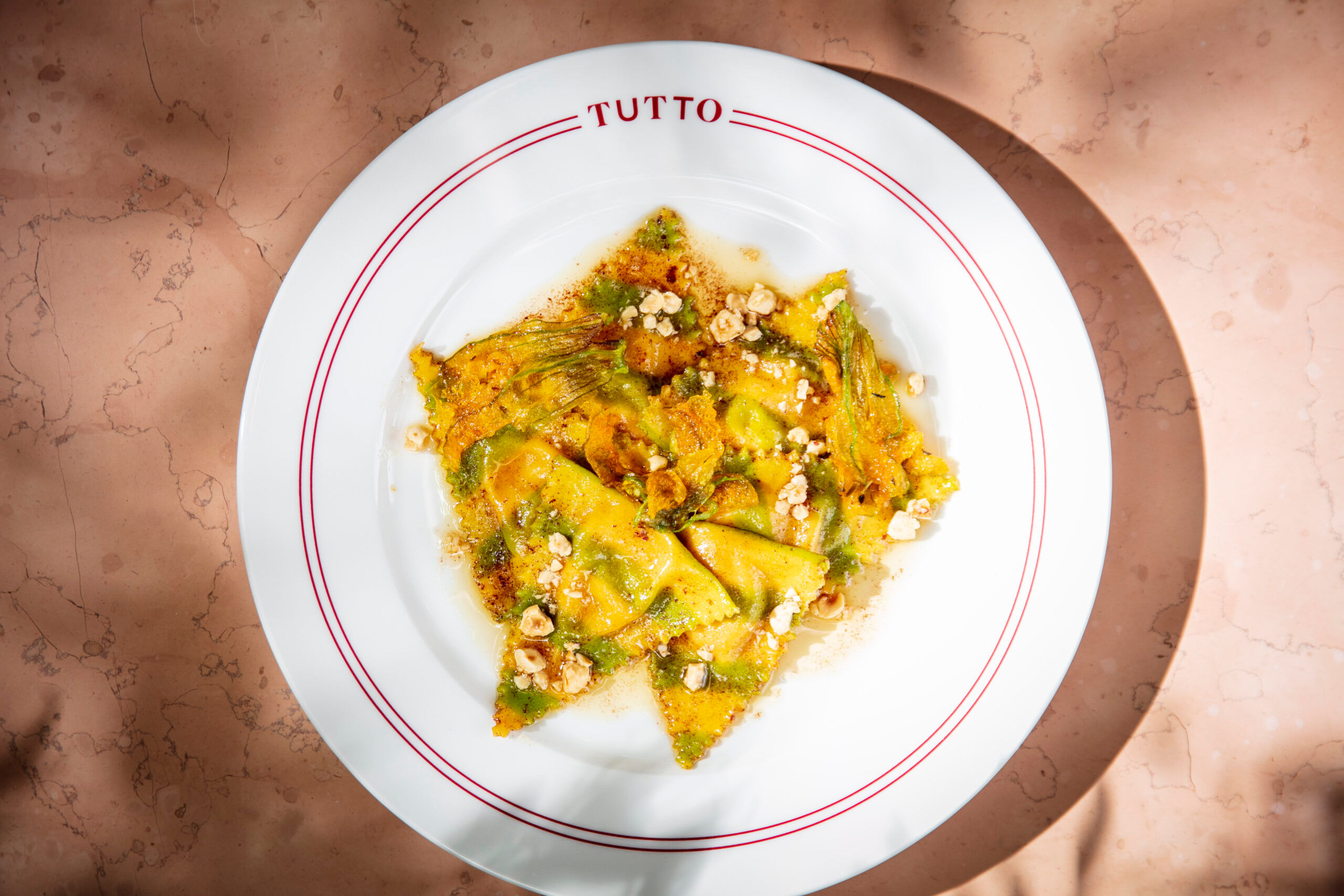 A white pasta dish photographed from above, around the rim is a double red line and the word TUTTO. The dish contains triangular filled pasta, wilted greens and crumbled nuts