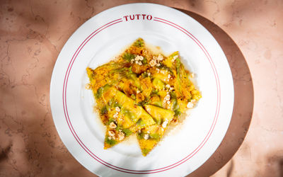 A white pasta dish photographed from above, around the rim is a double red line and the word TUTTO. The dish contains triangular filled pasta, wilted greens and crumbled nuts