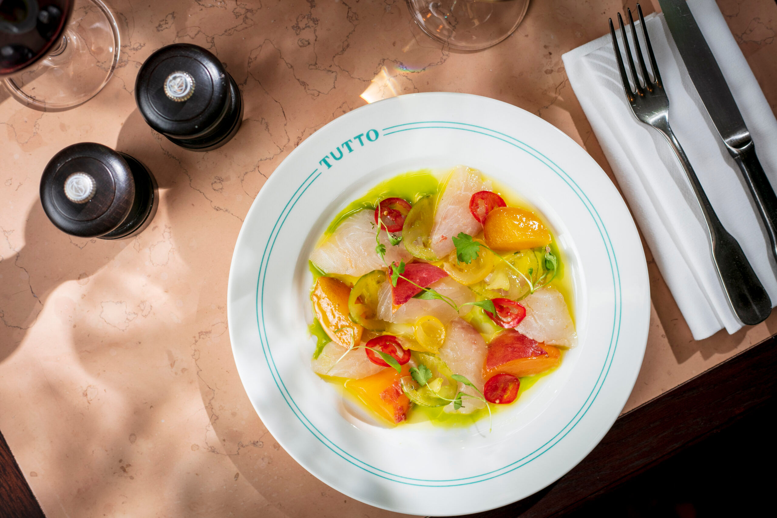 A white dish seen overhead with cutlery to its right and salt and pepper to the left. The dish contains fish, heritage tomatoes and stoned fruit in a green and yellow sauce