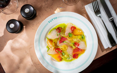 A white dish seen overhead with cutlery to its right and salt and pepper to the left. The dish contains fish, heritage tomatoes and stoned fruit in a green and yellow sauce