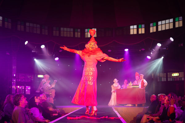 A drag queen in a red dress with red pointed hat with a heart on top is wearing clown makeup and stood with their arms held wide. In the background sits a panel of judges all smiling