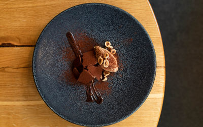 A slate grey flecked plate on the edge of a round wooden table holds a dessert dish of a deconstructed banoffee pie, a streak of chocolate sauce, crumbled