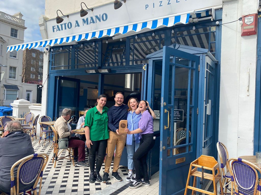 Four people are holding a wooden trophy outside a blue fronted pizzeria with a black and white tiled floor and blue and white striped awning.