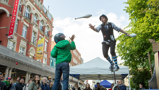 A crowd of onlookers surround a man on a tall unicycle as a child in a green jecket throws up a white juggling club to him