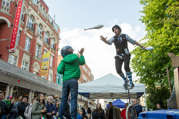A crowd of onlookers surround a man on a tall unicycle as a child in a green jecket throws up a white juggling club to him