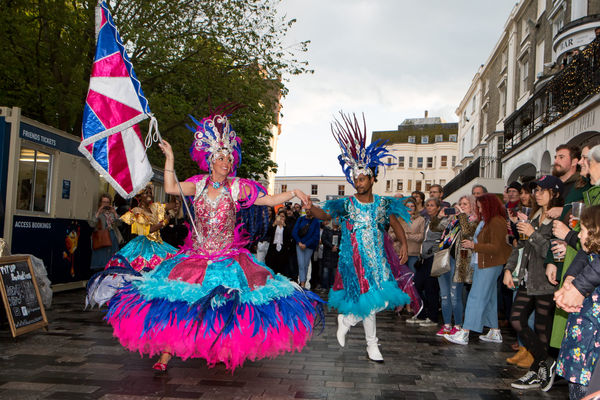 A woman is wearing a wide carnival dress in hot pink and electric blue, she has a feathered silver headdress and carries a pink and blue satine union flag in her right hand.She's holding hands with a man to her left who is wearing an electric blue carnival coat and feathered headdress. The couple are dancing though a street with crowds of onlookers about them.
