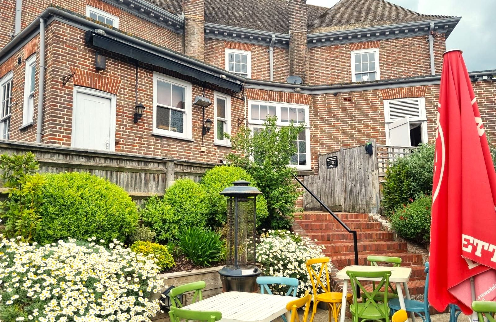 exterior shot of Ladies Mile pub, brick house with beautiful full of plants garden, white tables and chairs in different colors such as green, yellow, blue. There are couple of parasols