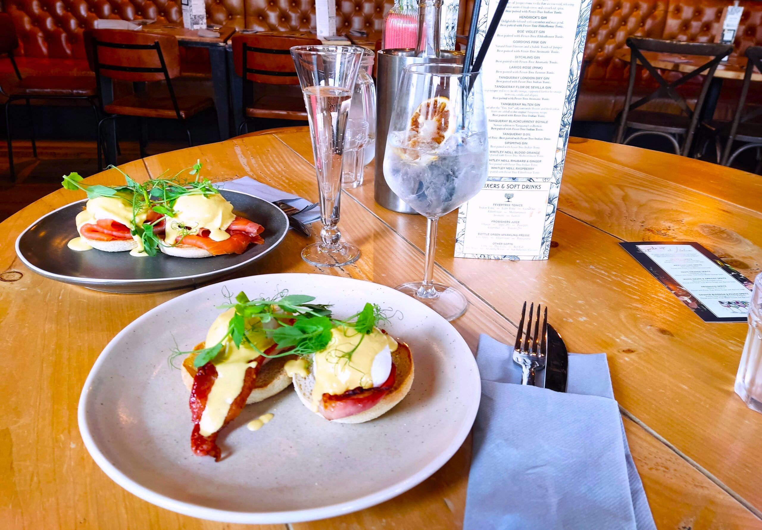 two white plates with egg hollandaise on the brown table together with some bottle of alcohol drink and two glasses