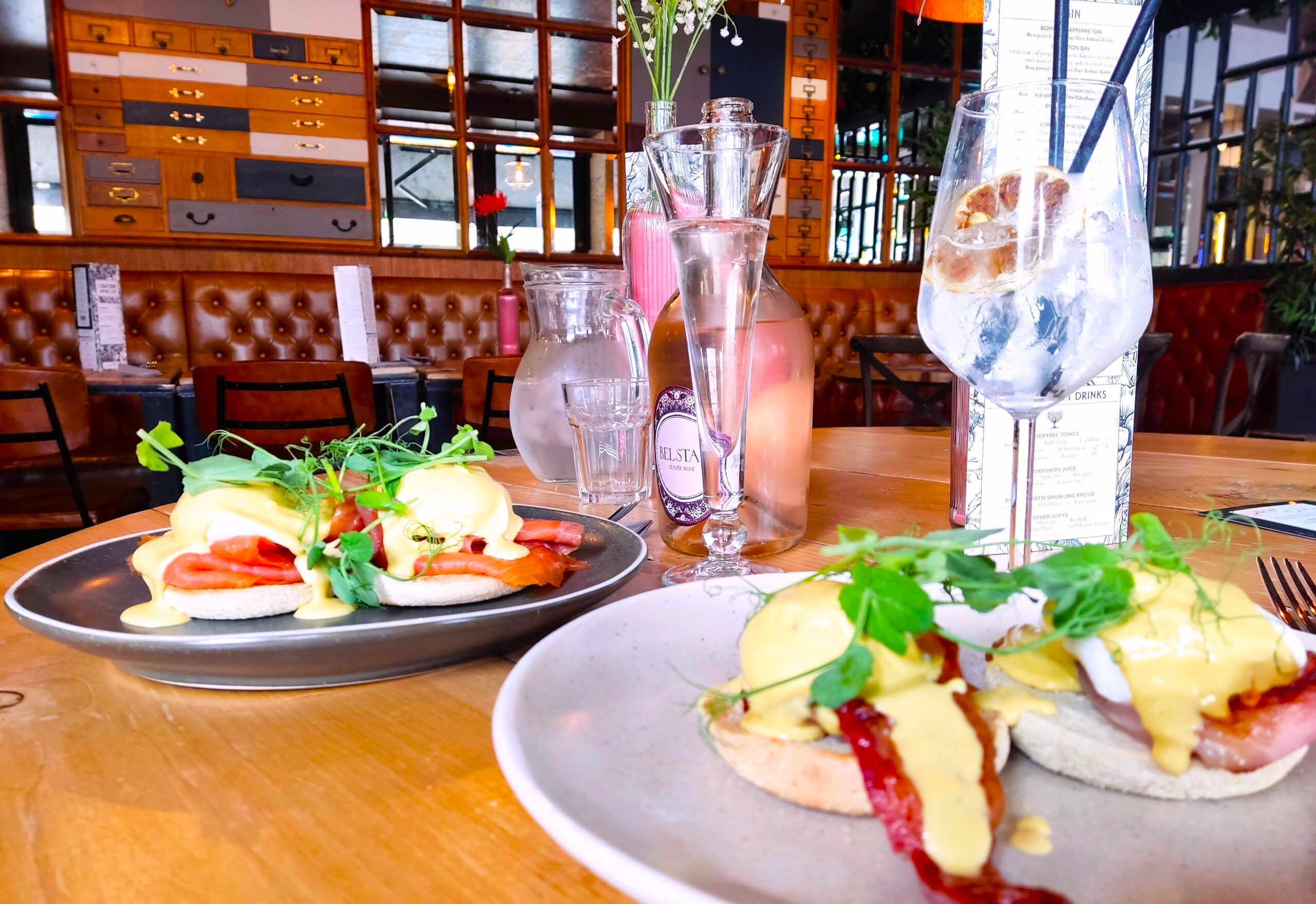 two white plates with egg hollandaise on the brown table together with some bottle of alcohol drink and two glasses. brunch at Libation bar 