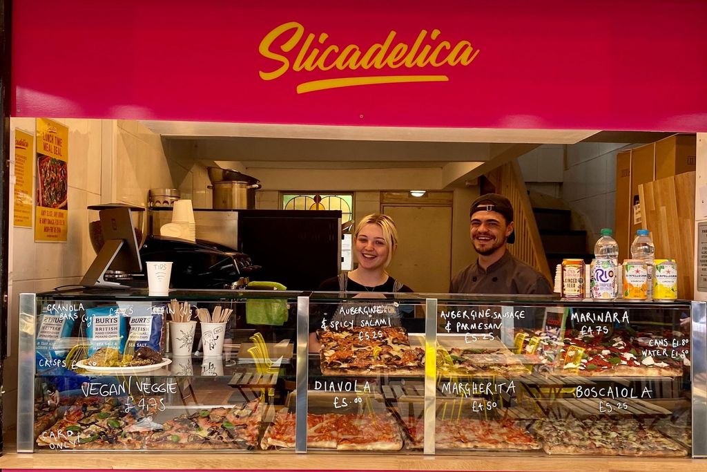 A man and a woman are inside a small kitchen behind a counter displaying pizzas, soft drinks and snacks. Above them there is a sign that is red with yellow writing saying Slicadelica
