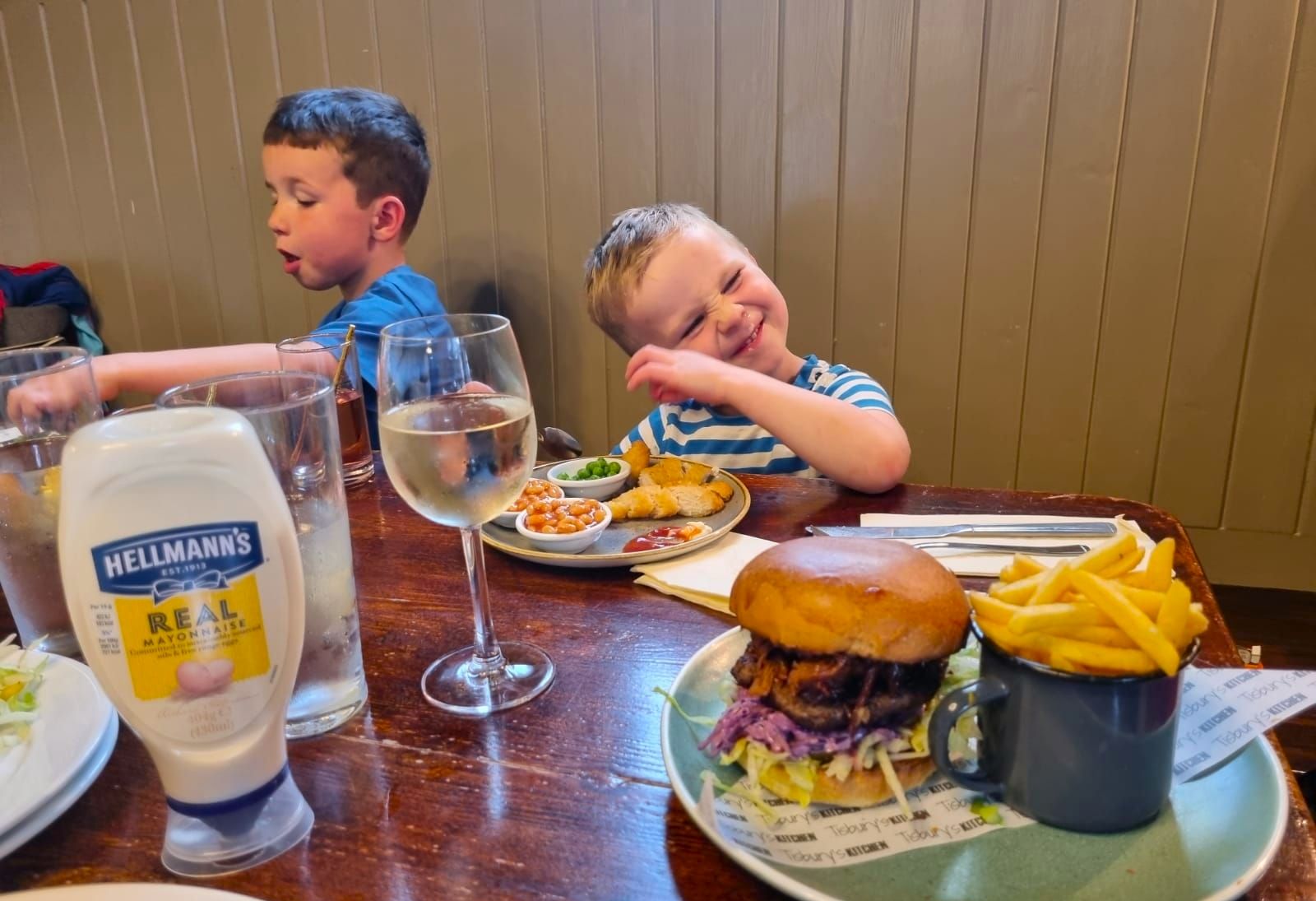 two kids enjoying their burger and fries at the table, their is glass of water and bottle of mayo on the table too