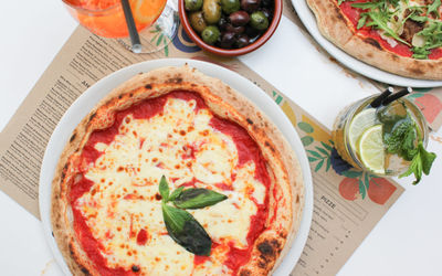 overhead shot of amargarita pizza topped with plenty of cheese and a sprig of basil. An Aperol and mojito as well as some mixed olives are also in view