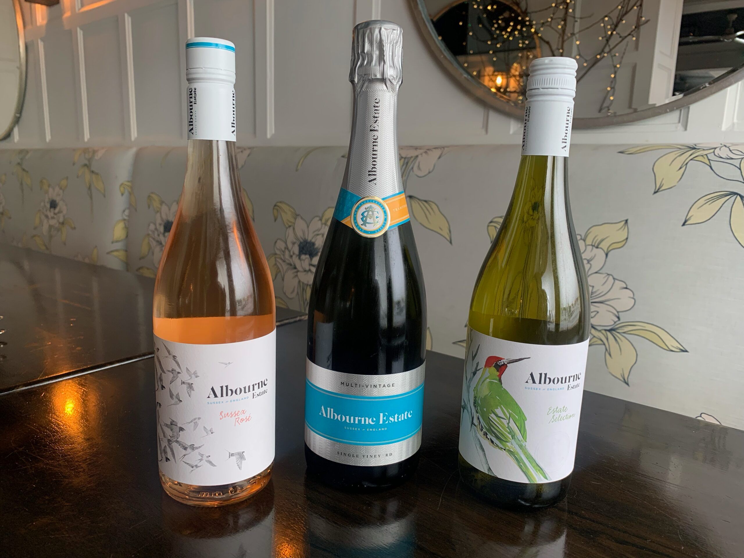 Three bottles of wine from the Albourne Estate in Sussex on a wooden table with floral patterned seating behind it. To the left is a bottle of rosé, centre is a bottle of sparkling with silver and blue foil and label, to the right is a green glass bottle of whinte wine with a red woodpecker on the label.