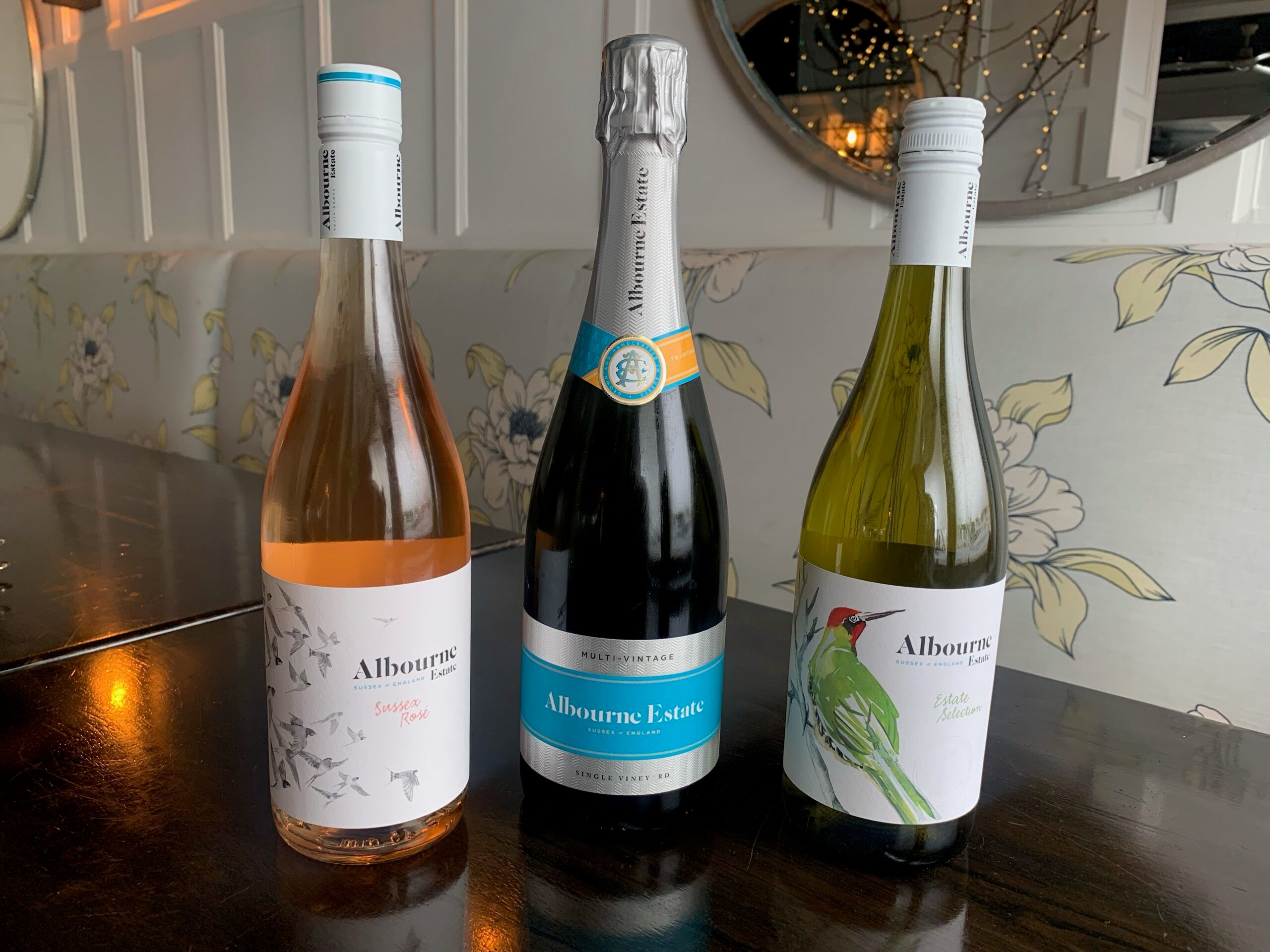 Three bottles of wine from the Albourne Estate in Sussex on a wooden table with floral patterned seating behind it. To the left is a bottle of rosé, centre is a bottle of sparkling with silver and blue foil and label, to the right is a green glass bottle of white wine with a red woodpecker on the label. Live Music Brighton blog