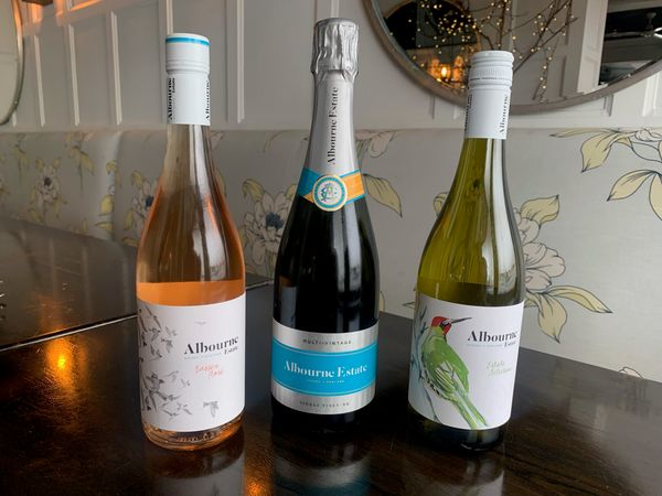 Three bottles of wine from the Albourne Estate in Sussex on a wooden table with floral patterned seating behind it. To the left is a bottle of rosé, centre is a bottle of sparkling with silver and blue foil and label, to the right is a green glass bottle of whinte wine with a red woodpecker on the label. 