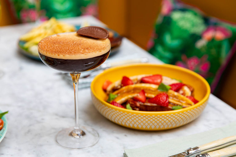 Espresso Martini cocktail in a tall glass with a yellow bowl of fresh fruit on a marble table.