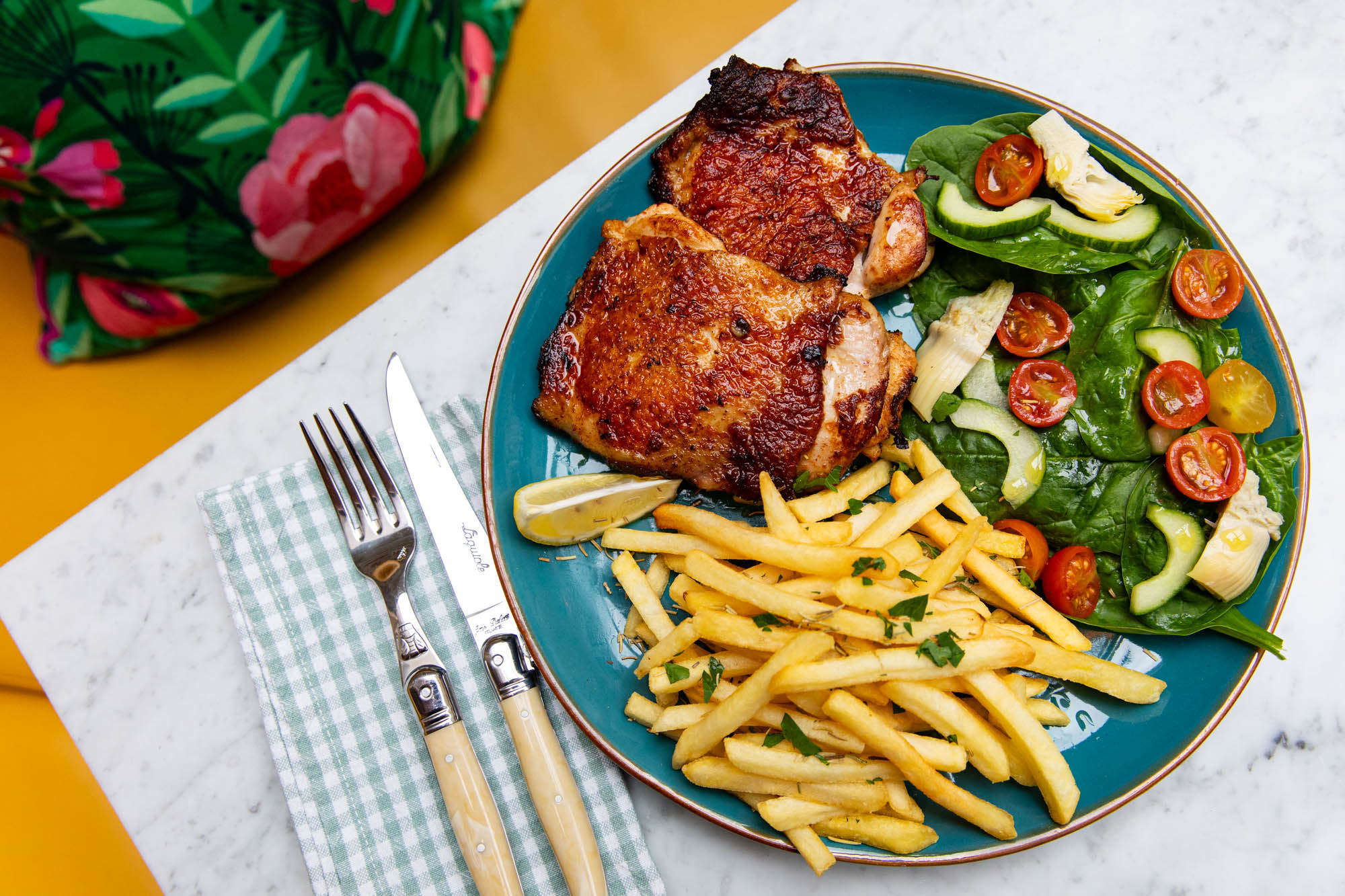 A plate of food on a blue plate with fries and a salad. A napkin with a knife and fork is positioned next to the food. Dinners at Jo and Co