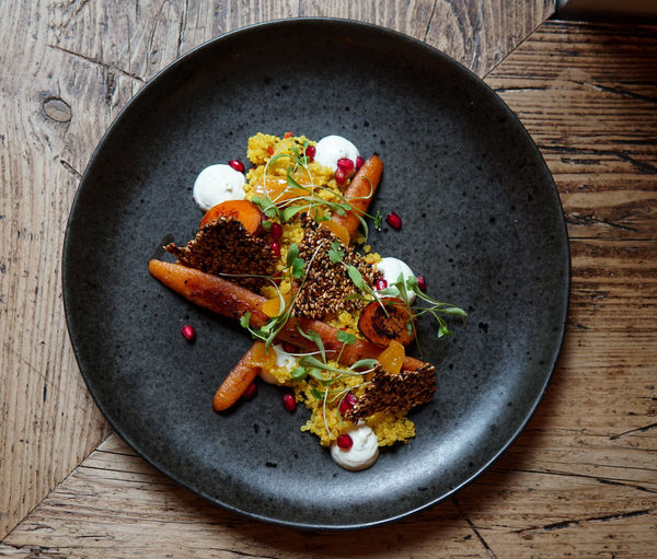 Overhead shot of a dark plate on a wooden table. Roasted carrots cut length ways on a bed of yellow couscous sesame seed brittle and pomegranate seeds
