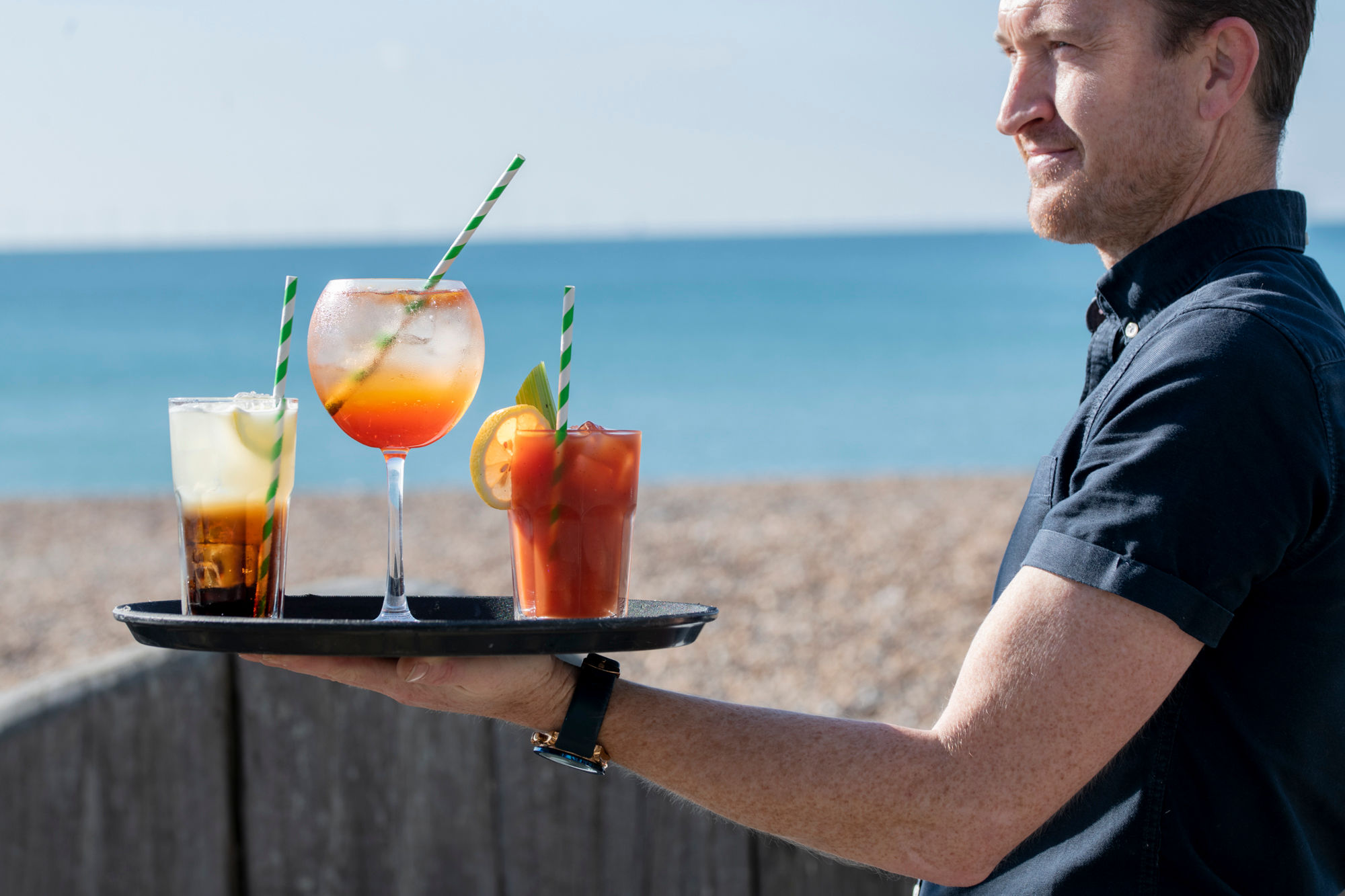 Man holding a tray with 3 cocktail glasses. Beach and sea in the background.