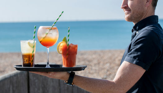 Man holding a tray with 3 cocktail glasses. Beach and sea in the background. Cocktail bars Brighton guide
