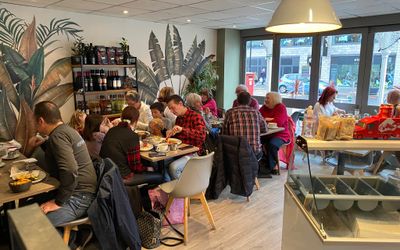 A view inside a cafe which is light with large windows and lots of plants, families are eating and all the tables are full, an ice cream cabinet is visible in the right of the picture